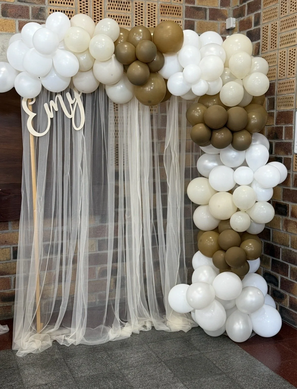 Décoration de baptême avec arche de ballons blanc, beige et doré, rideaux en voilage, prénom personnalisé en bois “Emy” et coin photo élégant réalisé par Les Jolis Instants.