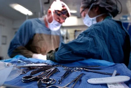 Two surgeons in scrubs and masks performing surgery in an operating room with surgical instruments on a table in front of them.