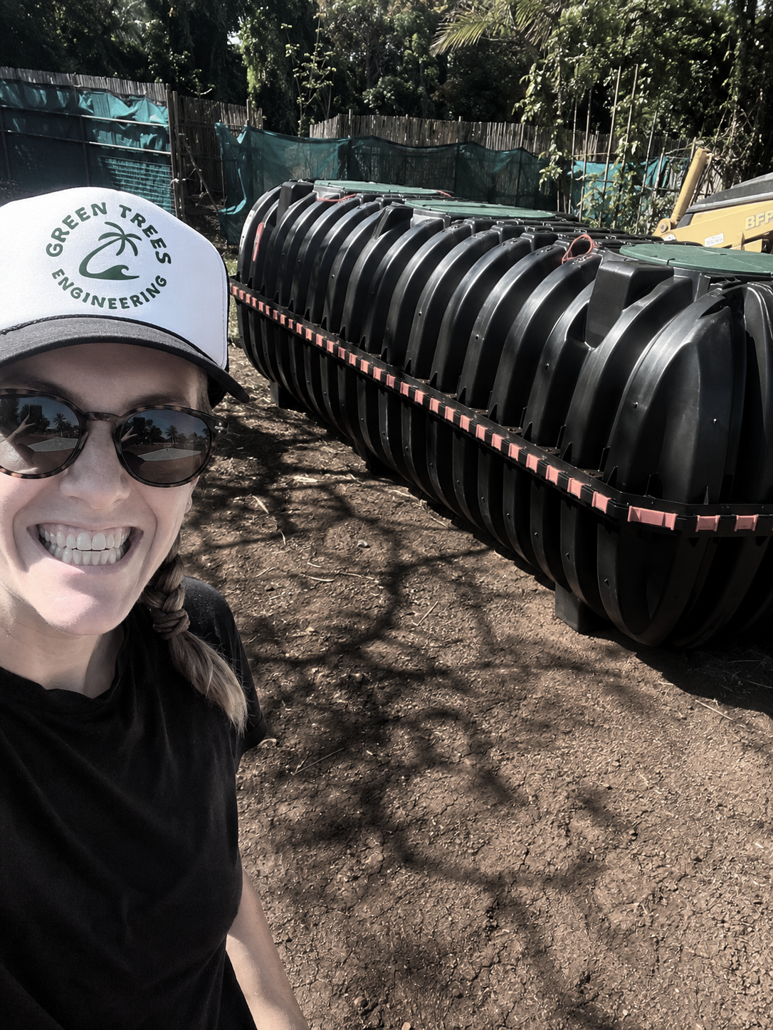 A smiling woman wearing sunglasses and a cap with the Green Trees Engineering logo taking a selfie outdoors near a large black plastic composting or waste-processing container, with trees and fencing in the background.