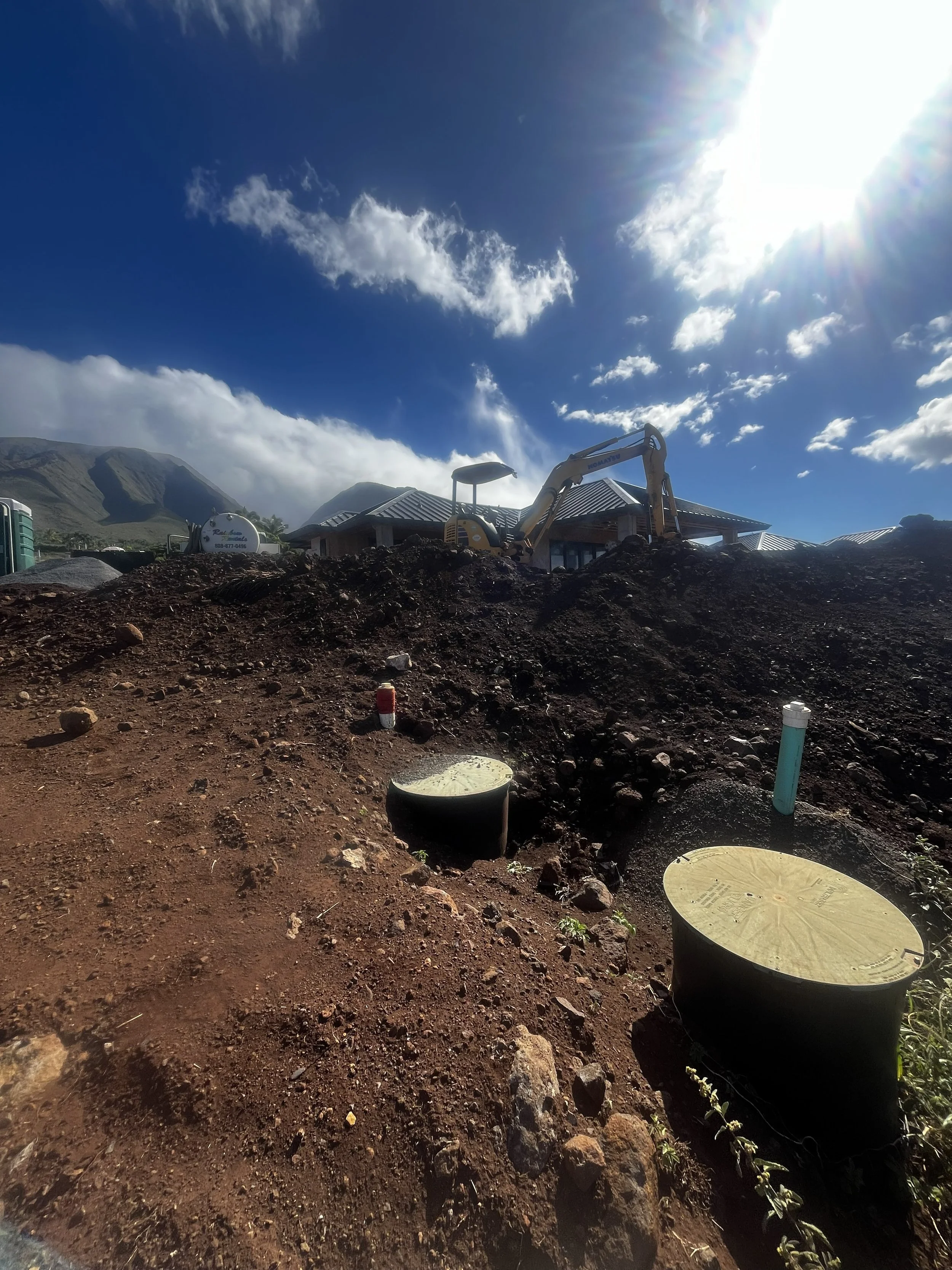 Construction site with a small excavator, construction materials, and pipes in a mountainous area under a bright blue sky with scattered clouds and the sun shining.