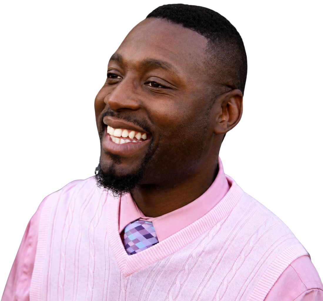 A smiling man with a short beard and mustache, wearing a pink sweater over a pink dress shirt and a purple patterned tie.