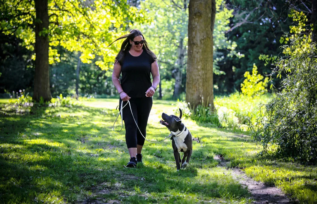A woman with dark hair, wearing sunglasses, a black t-shirt, and black leggings, is walking a dog on a leash in a sunny park with green trees and grass.