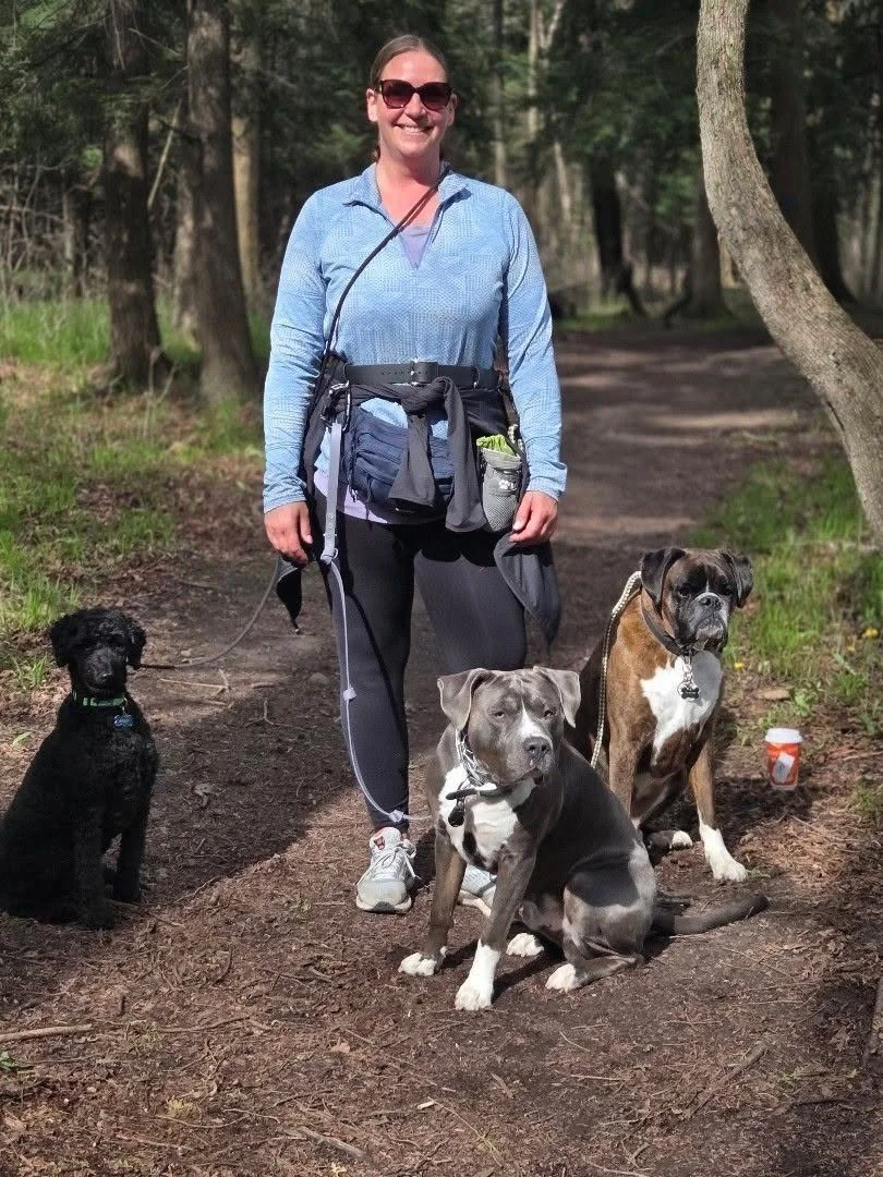 A woman standing on a dirt trail in a forest, smiling with three dogs sitting or standing nearby.
