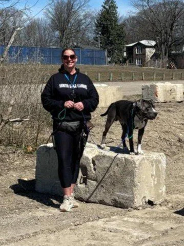 A woman in a black hoodie and sunglasses standing on a concrete block, holding a leash attached to a dog standing on the same block, outdoors with trees and a building in the background.