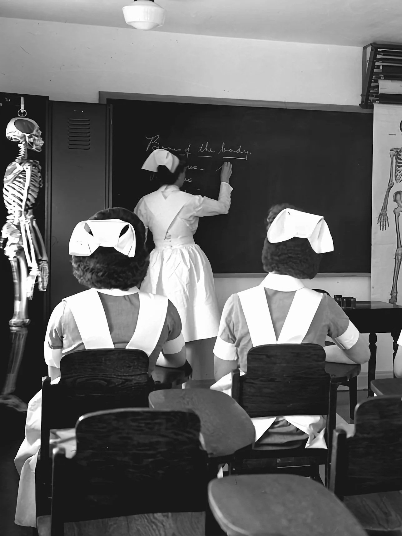 A classroom scene with women in vintage nurse uniforms, including hats, sitting at desks facing a blackboard where a woman is writing. The classroom has anatomical posters and a skeleton model, suggesting a medical or nursing educational setting.