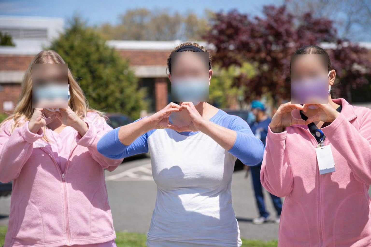 Three women outdoors making heart shapes with their hands, wearing masks, with trees and buildings in the background.