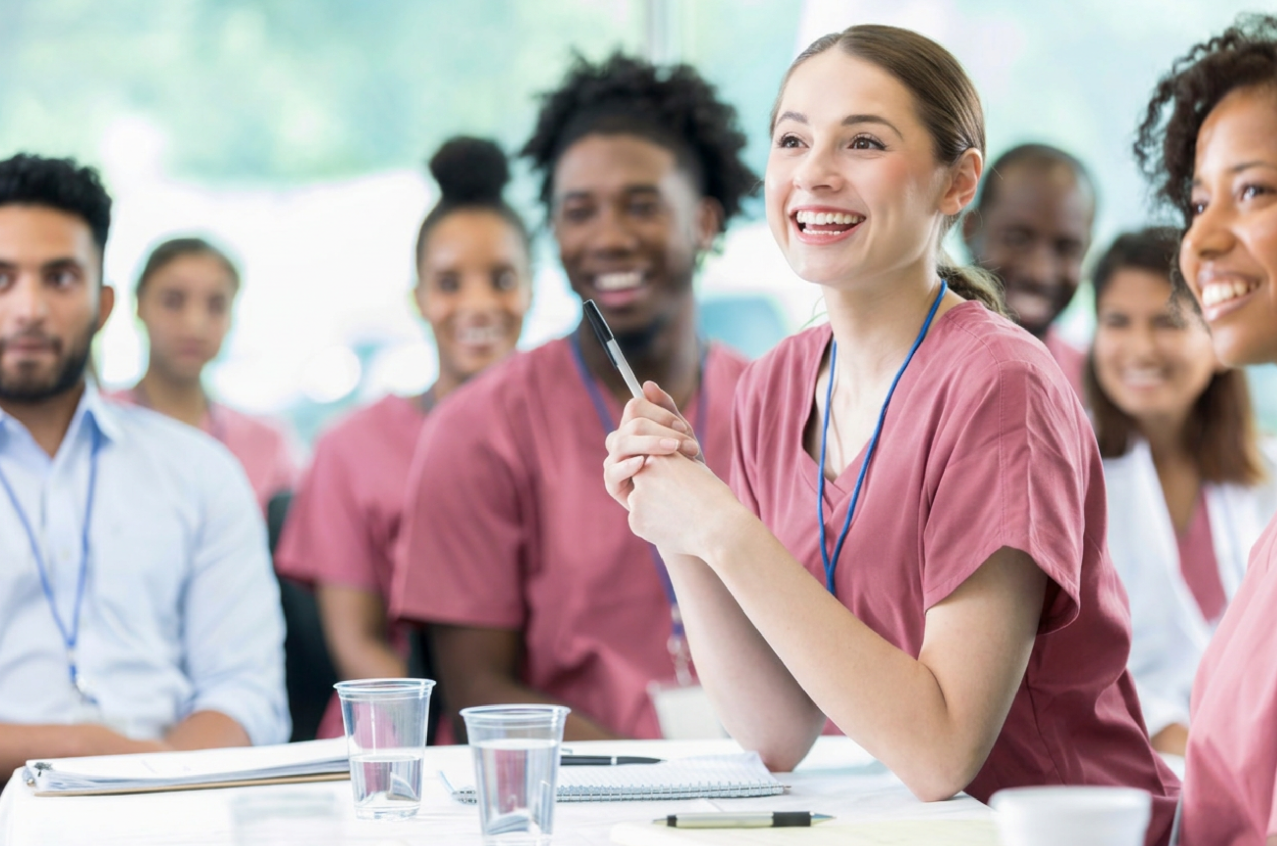 A group of diverse young adults in pink scrubs and casual clothing sitting around a table with drinks, smiling, and engaging in a discussion in a bright, modern setting.