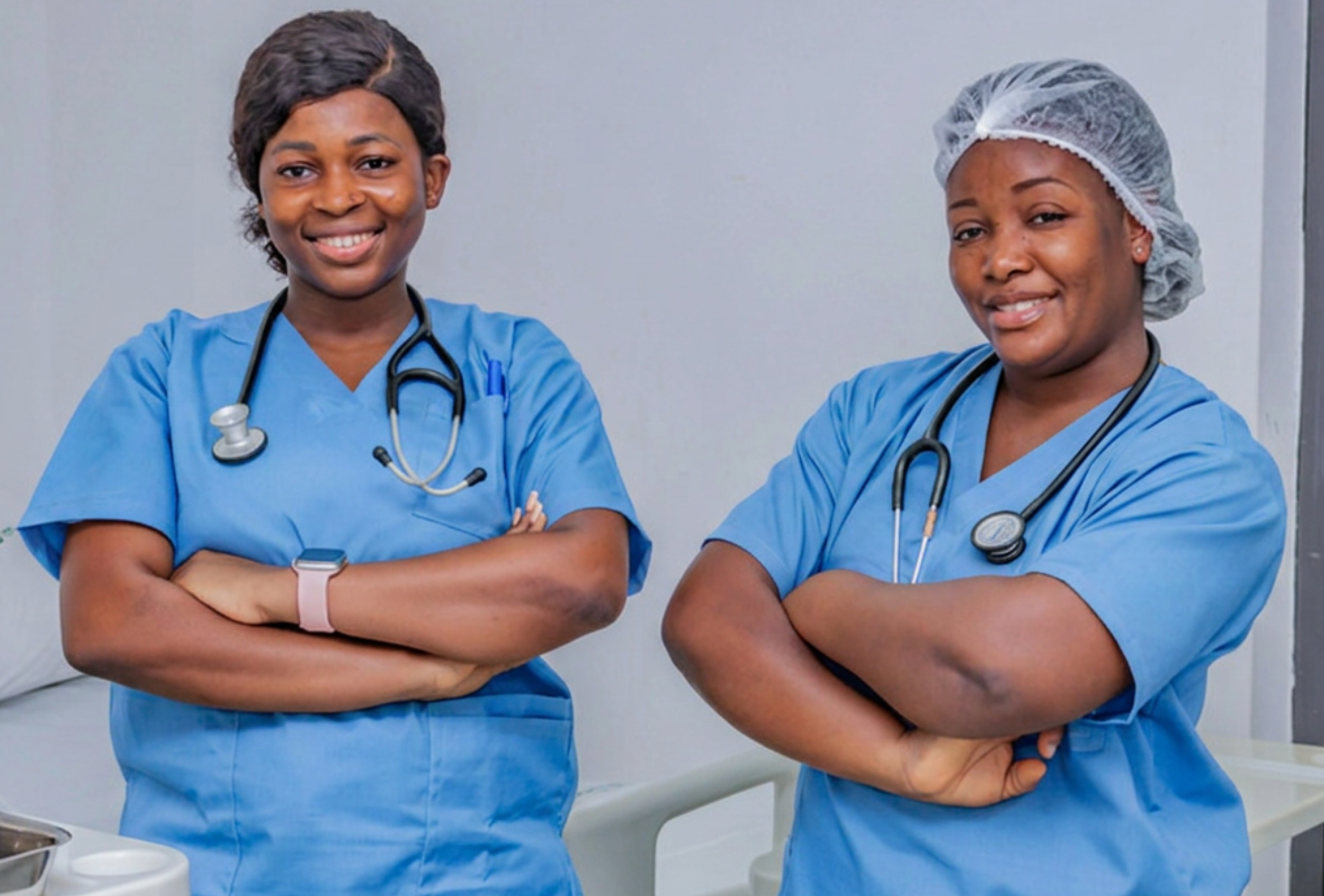 Two female healthcare workers in blue scrubs with stethoscopes standing with their arms crossed and smiling in a clinical setting.