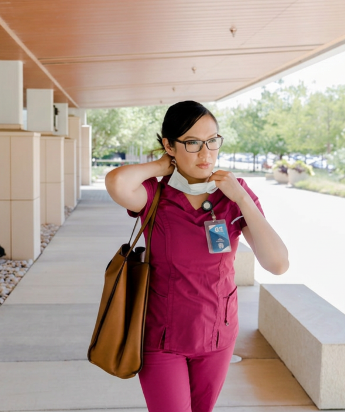 A female healthcare worker in pink scrubs adjusts her face mask outside a medical facility.