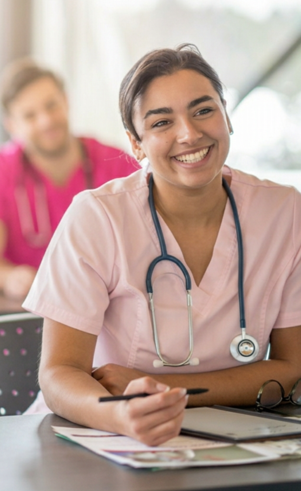 A smiling female nurse in pink scrubs with a stethoscope around her neck sitting at a desk with some papers and a pen.