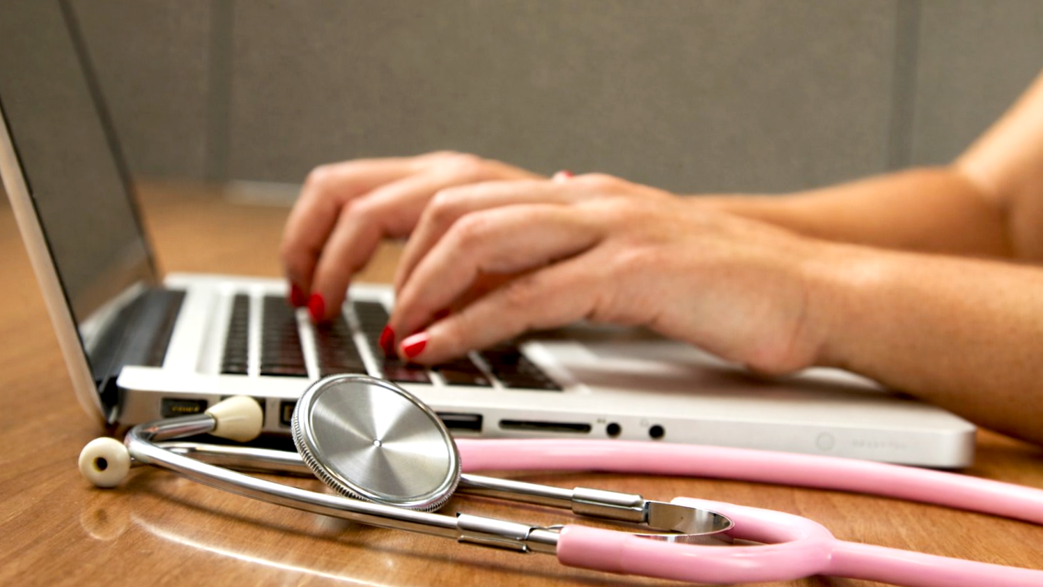 Close-up of female hands with red nails typing on a laptop keyboard, with a pink stethoscope resting on the wooden desk in the foreground.