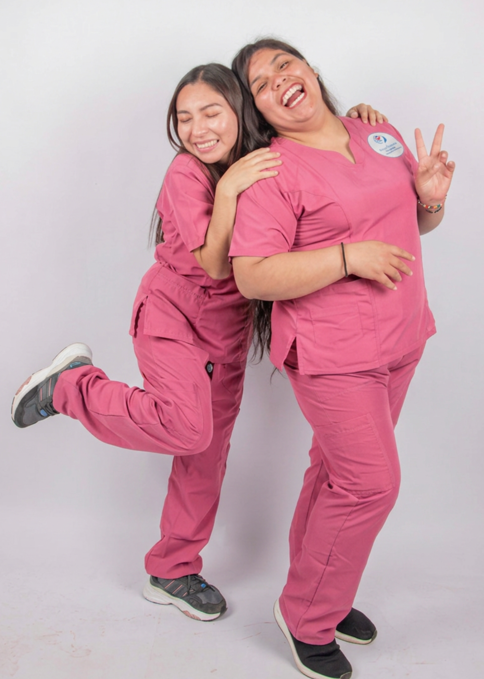 Two women in pink scrubs are smiling and having fun together, one giving a peace sign and the other hugging her from behind, standing against a plain white background.