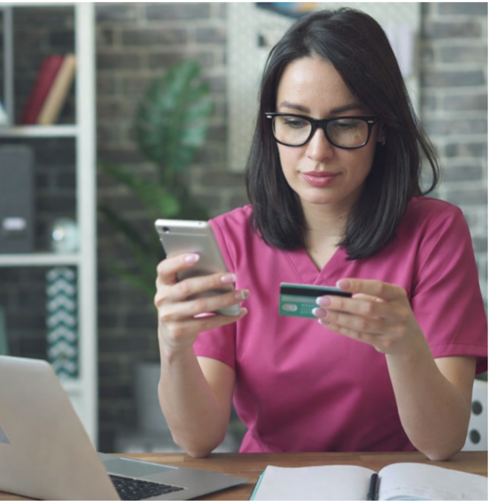A woman in pink scrubs sitting at a table, looking at her smartphone and holding a credit card, with a laptop and open notebook nearby.