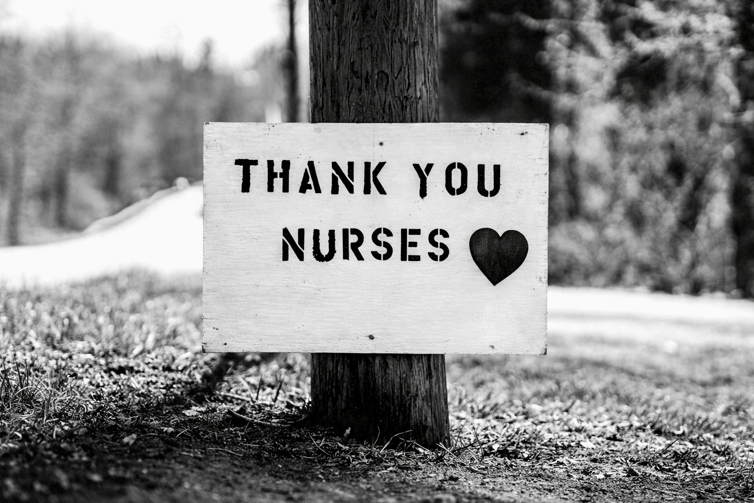 A wooden sign attached to a tree with the message 'Thank you nurses' and a black heart symbol, in a grassy outdoor area with trees in the background, in black and white.