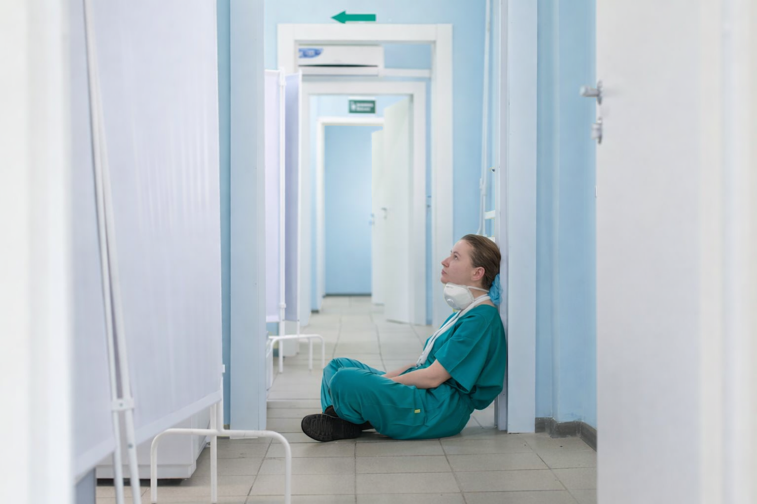 A healthcare worker in teal scrubs and a face mask sits against a light blue wall in a hospital hallway, looking to the side.