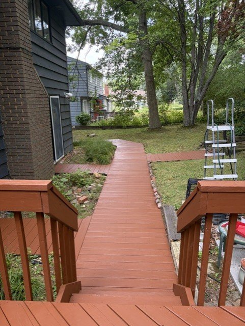 View of a backyard with a wooden deck stairs leading to a winding red wooden pathway, surrounded by green grass, trees, and shrubbery, with a ladder and some gardening tools on the right side.