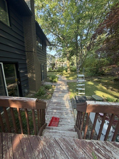 View of a backyard path seen from a wooden deck, with stairs leading down. A metal ladder is positioned on the grass, and trees and greenery surround the area.