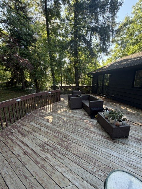 An outdoor wooden deck with wicker furniture and potted plants, surrounded by trees and a black house.