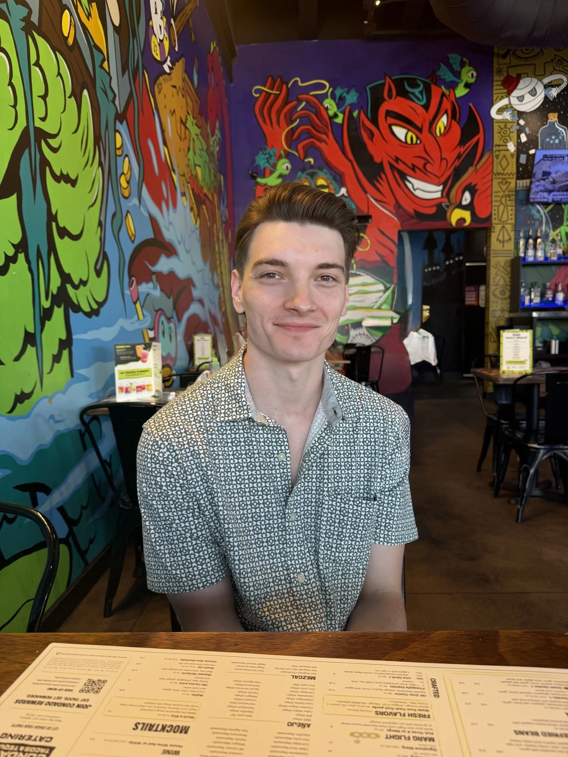 A young man with short brown hair, wearing a patterned short-sleeve shirt, sitting at a dining table in a restaurant with colorful graffiti-style murals on the walls featuring cartoon characters and vibrant colors.