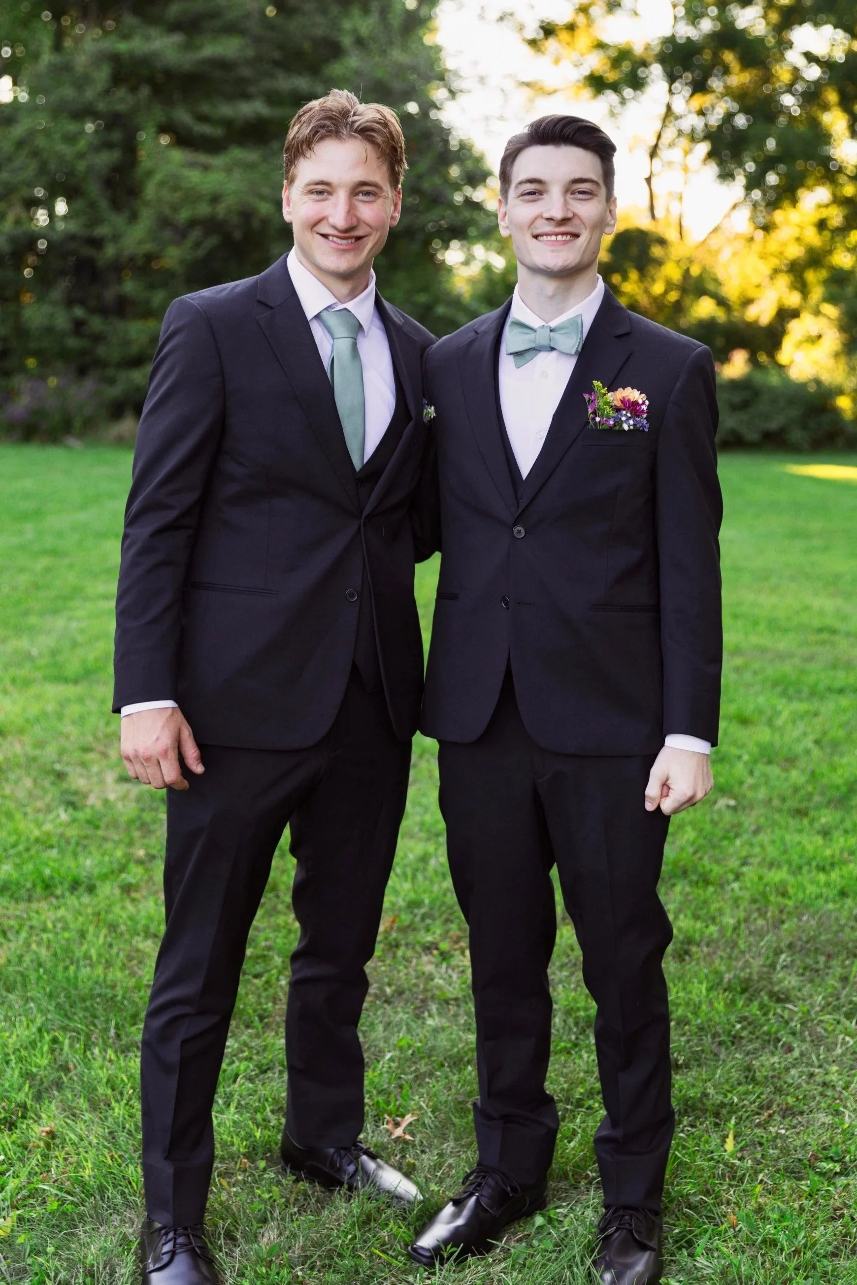 Two young men in formal suits and bow ties standing outdoors on grass with trees and sunlight in the background, smiling at the camera.