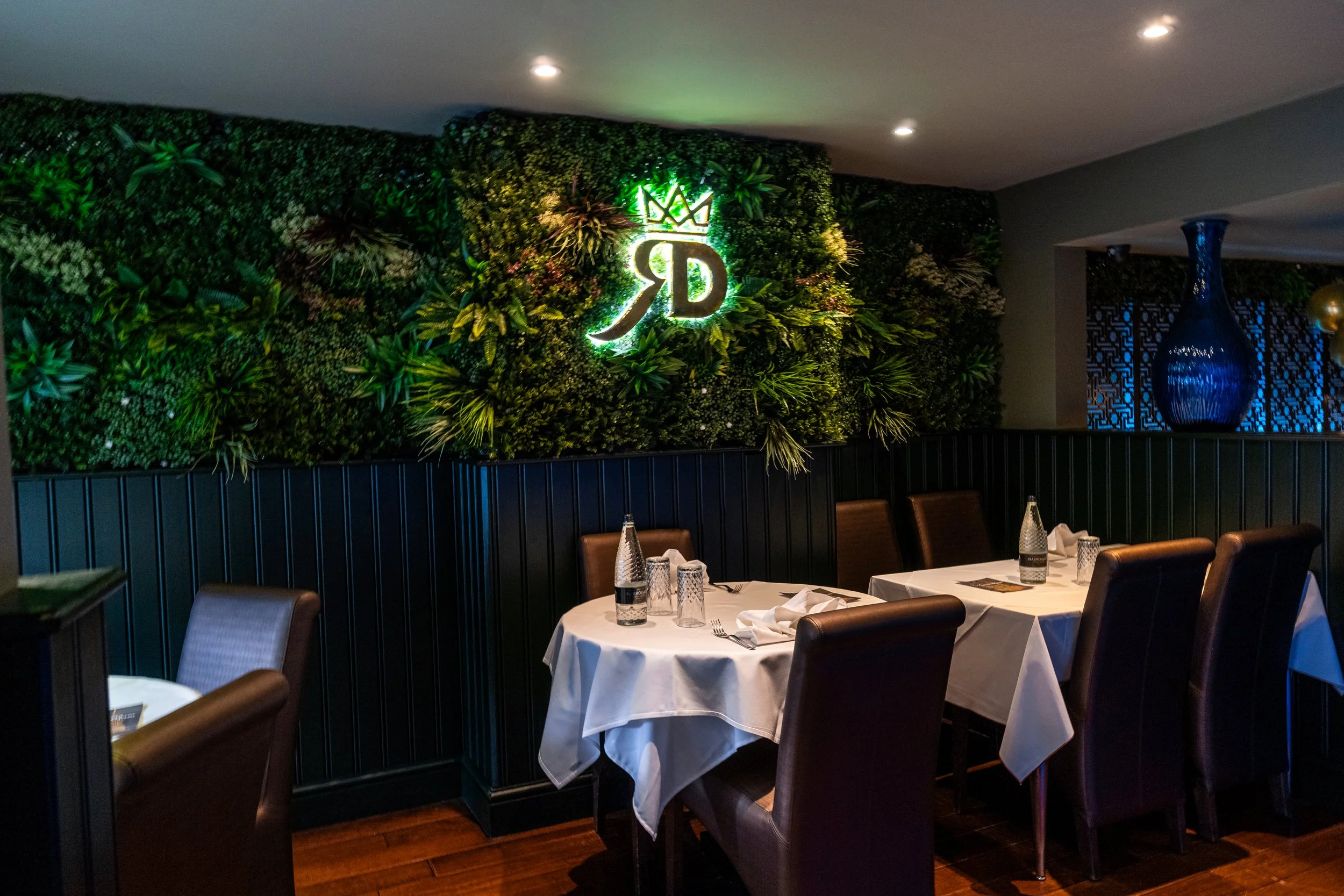 A restaurant dining area with tables covered in white tablecloths, set with glasses and napkins. The background features a decorative green wall with illuminated initials "RD" and a crown symbol, alongside a large blue vase and patterned wall decor.