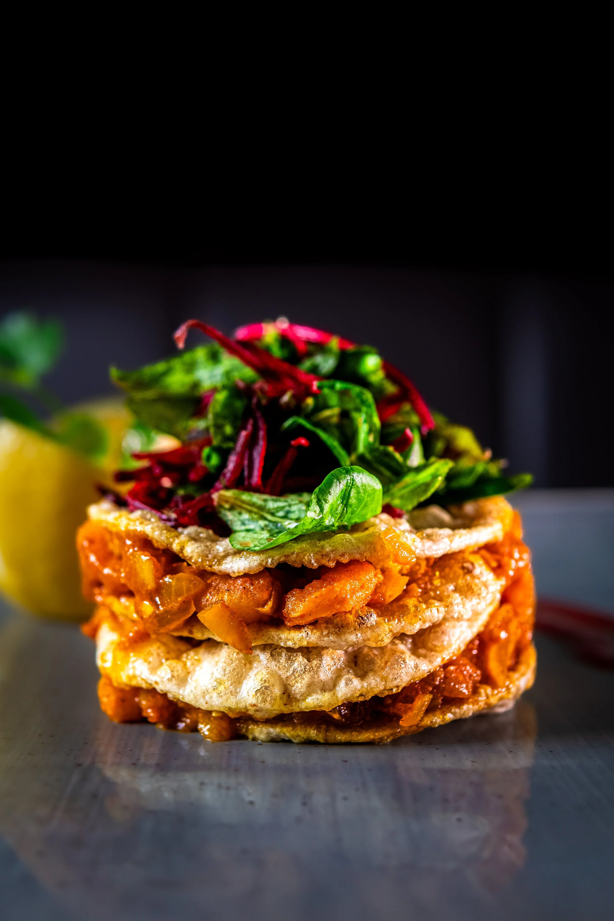 Close-up of a multi-layered appetizer with crispy base, topped with cooked minced vegetables, fried cheese, fresh greens, and microgreens, against a dark background.