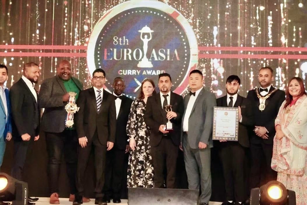 Group of people in formal attire celebrating at the 8th EuroAsia Curry Award event; some are holding awards and certificates, with a large screen displaying the event logo in the background.