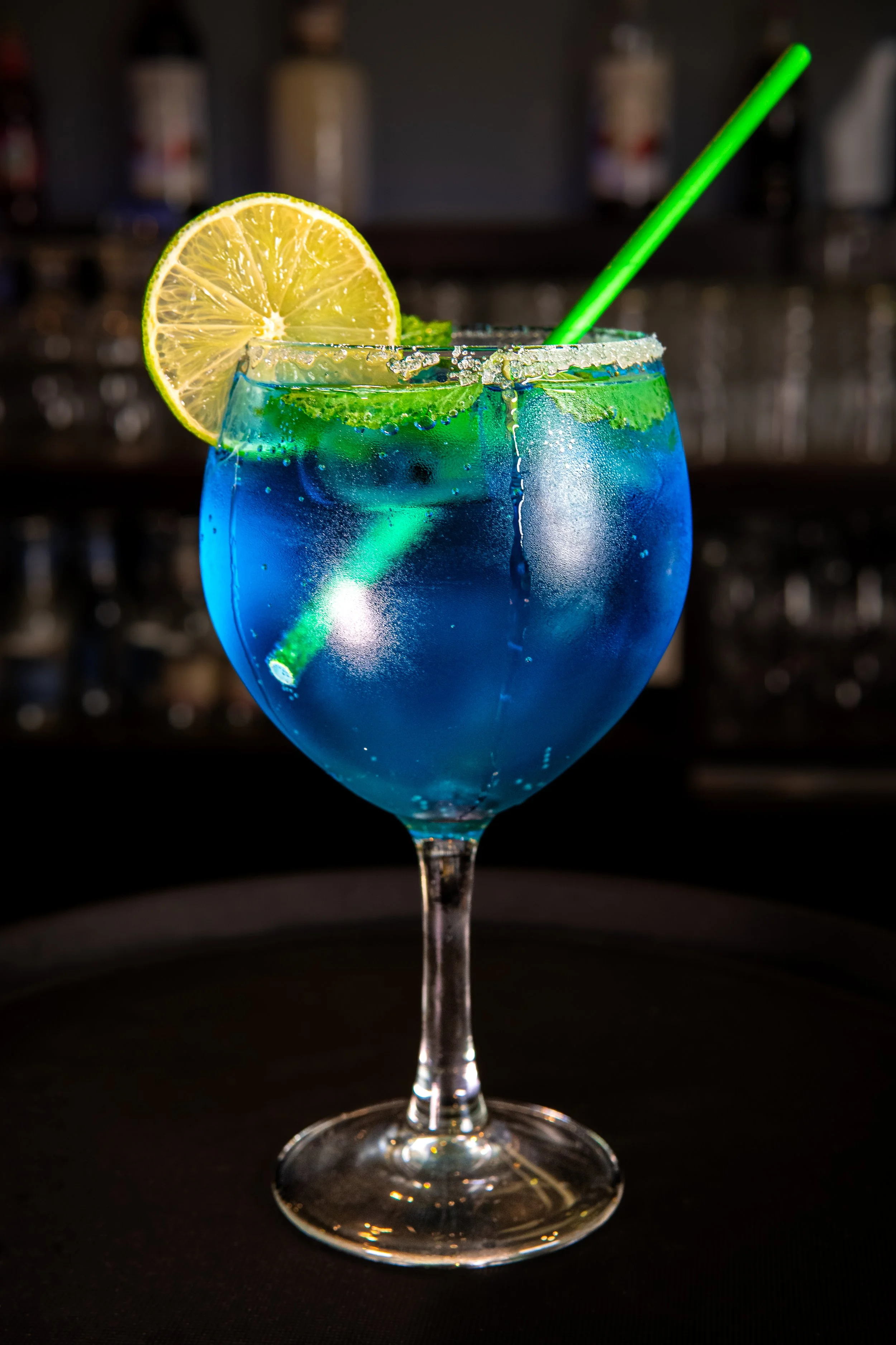 A colorful cocktail in a large wine glass with a salted rim, garnished with a slice of lemon and a green straw, set against a dark, blurred background.