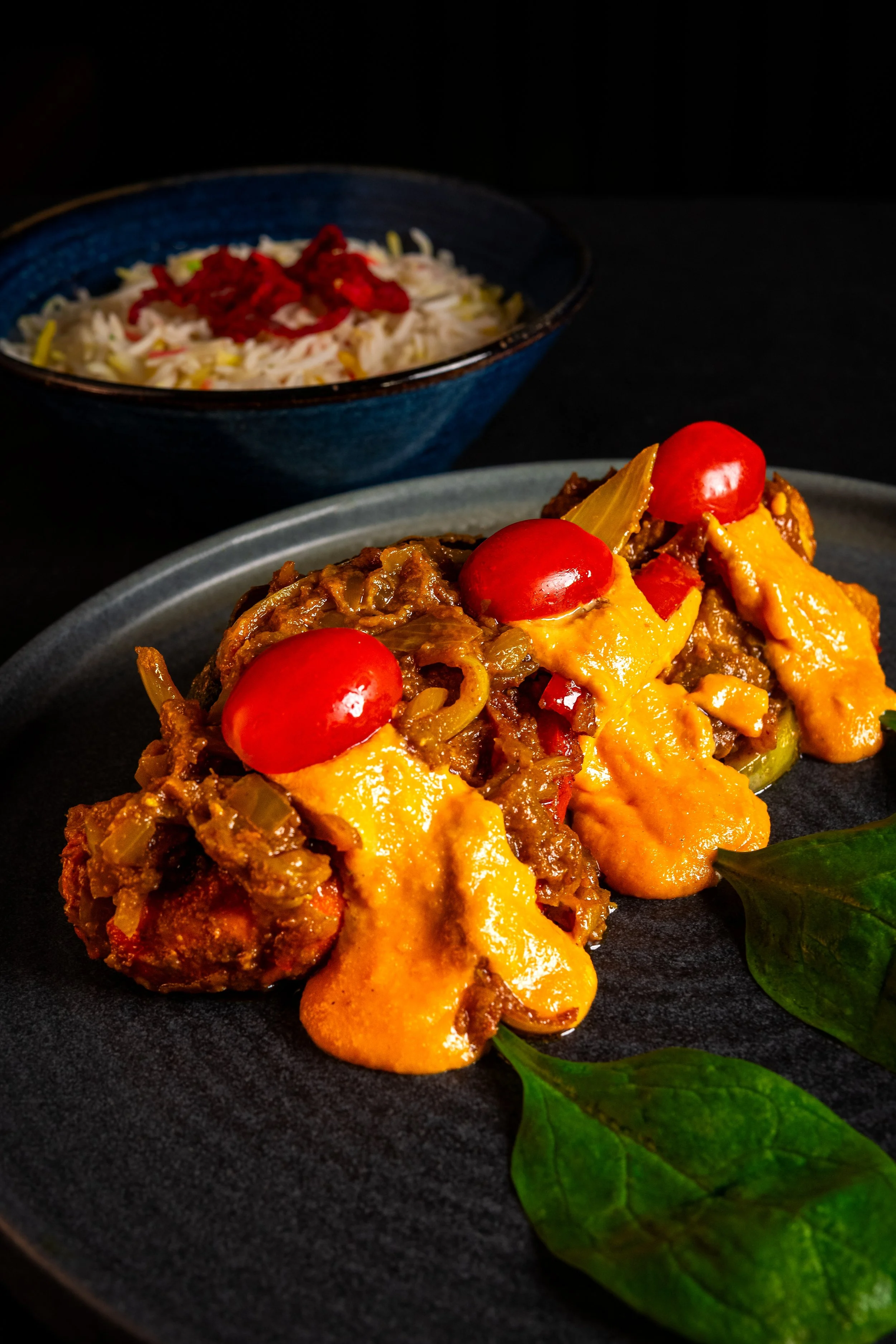 Close-up of a meatloaf with melted cheese, cherry tomatoes, and green herbs on a black plate, with a bowl of rice topped with red dried herbs in the background.