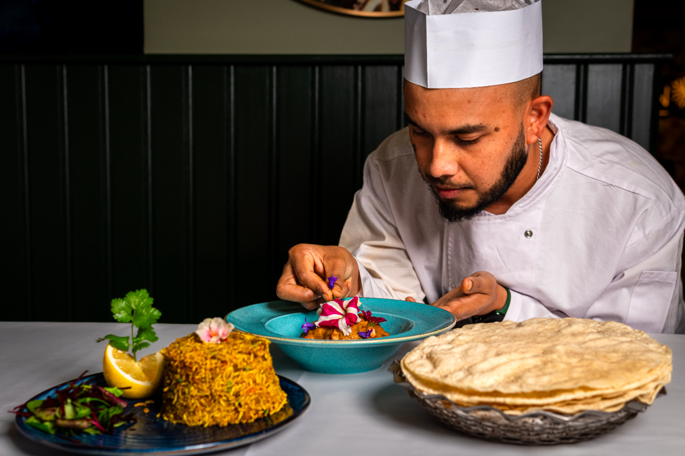 Chef in white uniform and hat carefully decorating a colorful dish with edible flowers at a restaurant table.