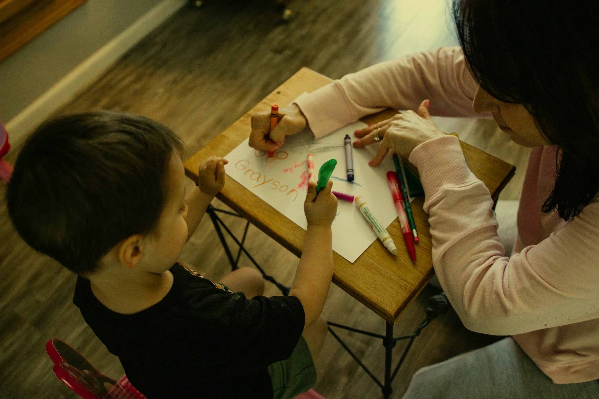 A woman and a young girl sitting at a wooden table, drawing on a piece of paper with colorful markers.