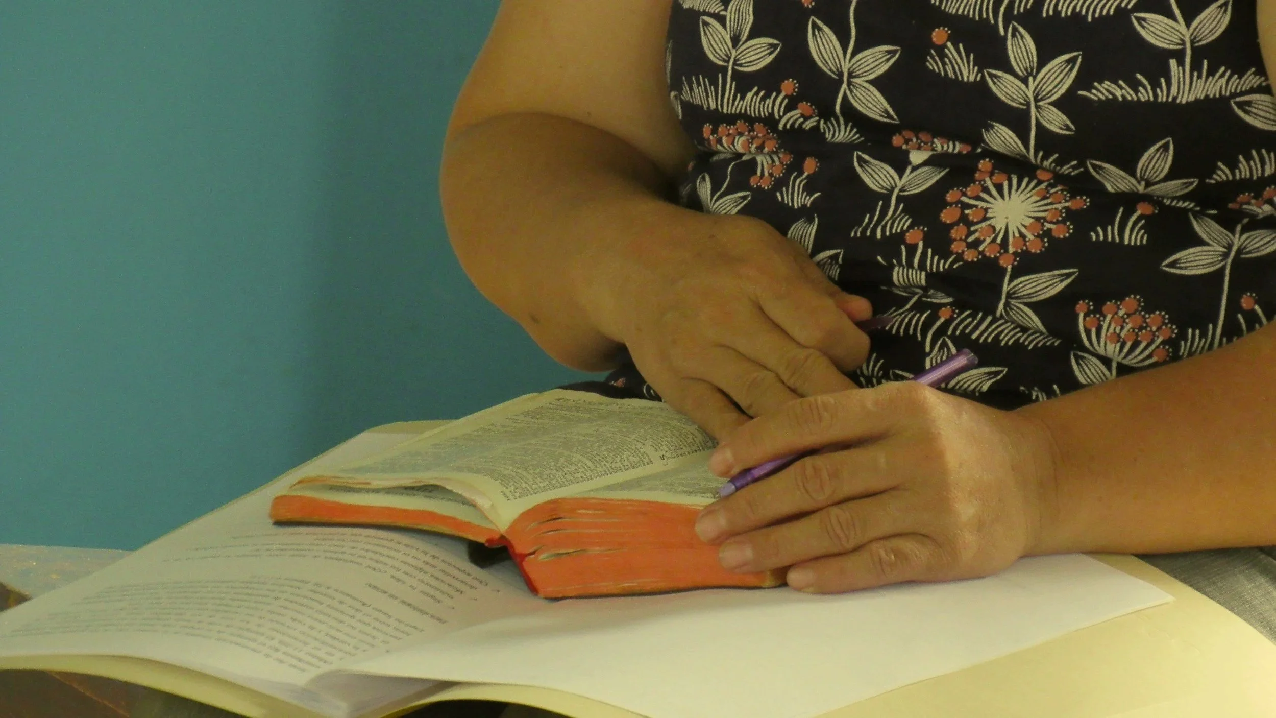 A person wearing a black floral patterned shirt is sitting at a table with open books, holding a purple pen, and writing or reading.