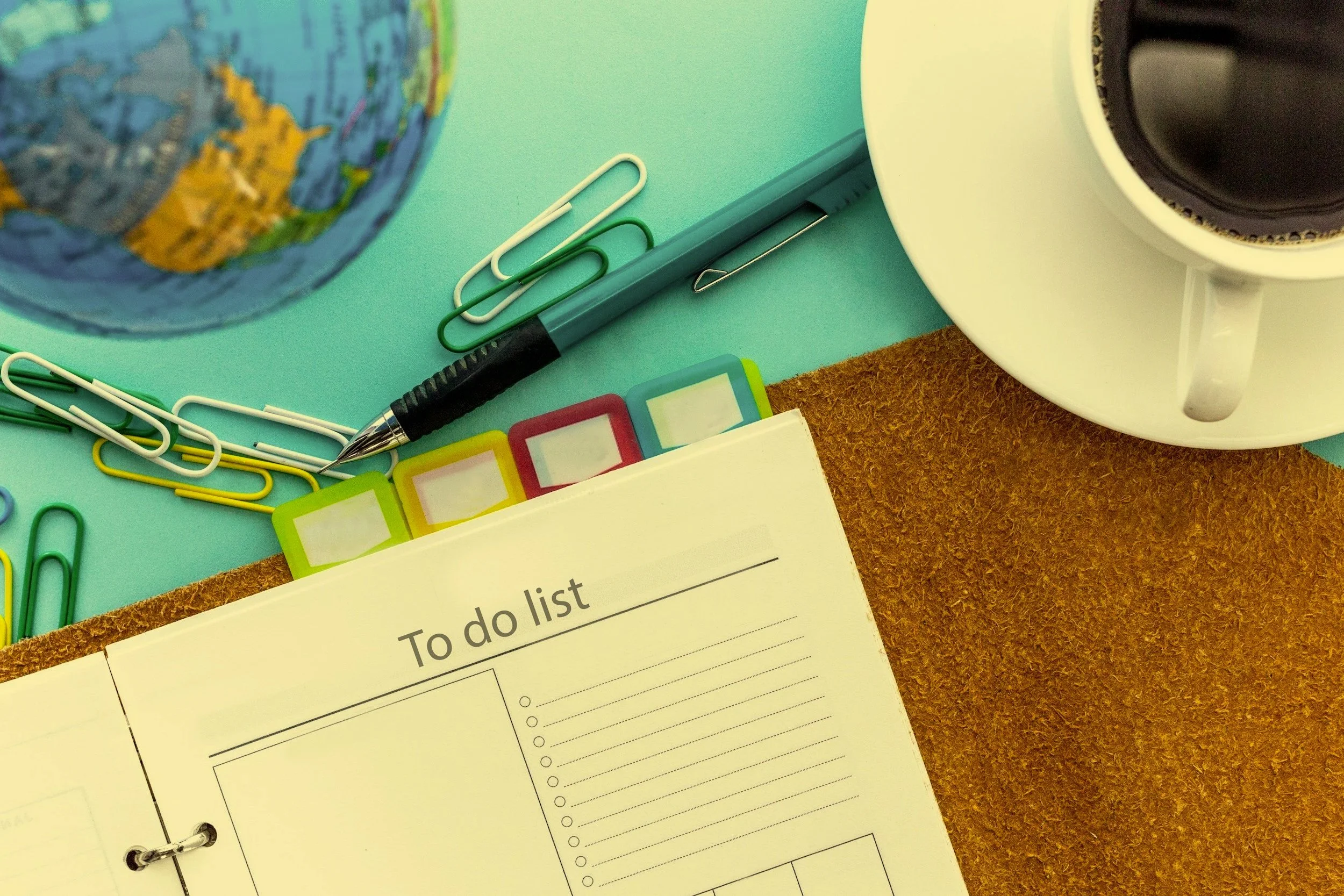 A turquoise sheet with a printed to-do list, colorful paper clips, a black and silver pen, a globe, and a white speaker on a brown surface.