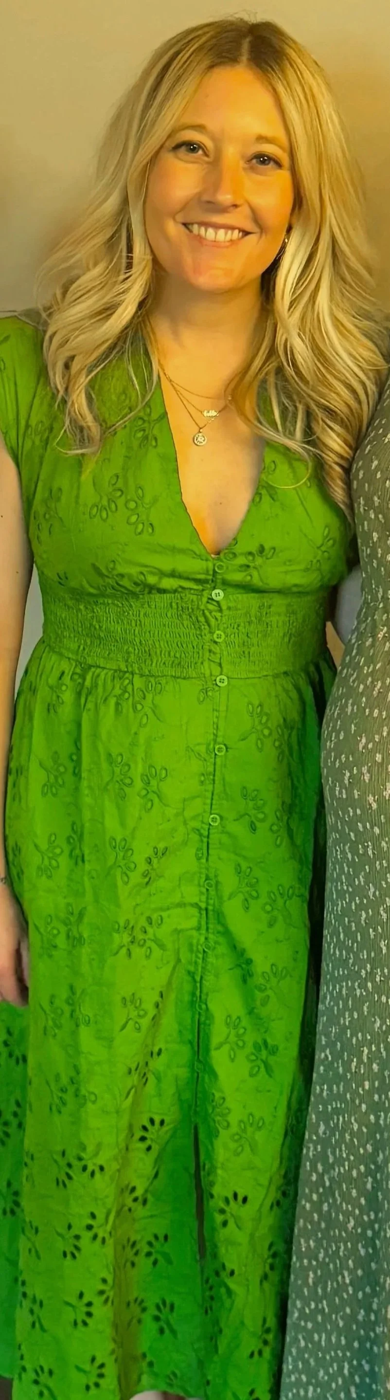 Smiling woman with long blonde hair in a green embroidered dress, standing indoors with a neutral background.