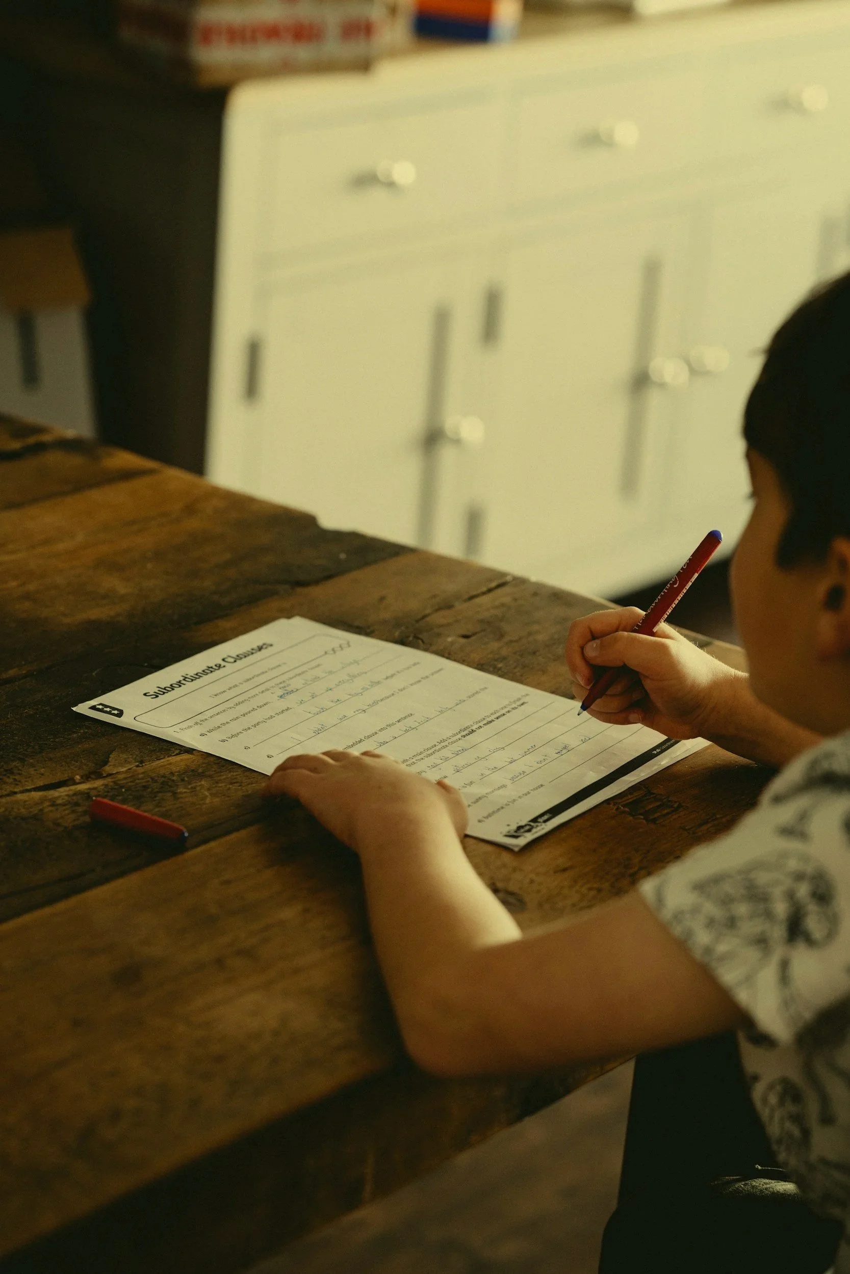 A child with dark hair doing homework or writing on a worksheet at a wooden table, with a white cabinet and some boxes in the background.