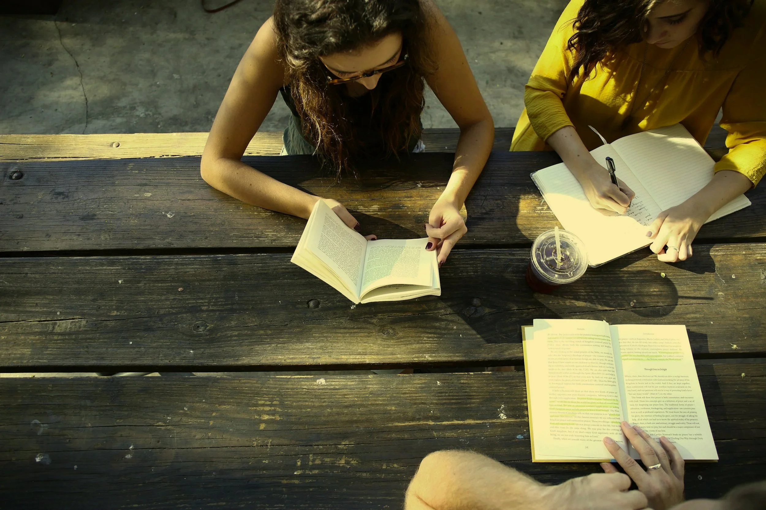 Two women sitting at a wooden picnic table, reading books on a sunny day, with one holding a notebook and pen, and the other holding an open book, and a large iced drink with a straw on the table.