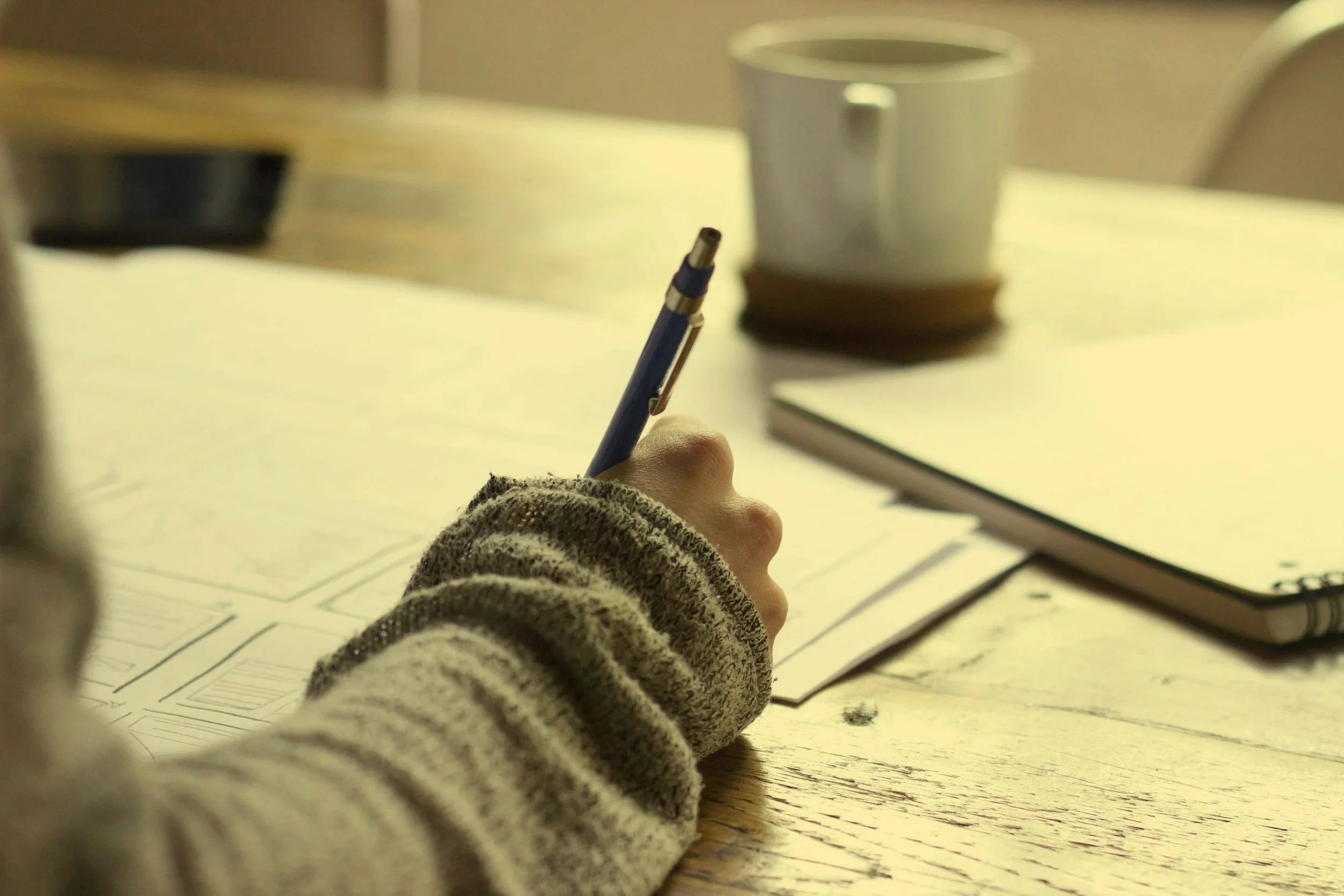 A person's hand holding a pen over paper with sketches or drawings, on a wooden table, with a coffee mug and notebook in the background.