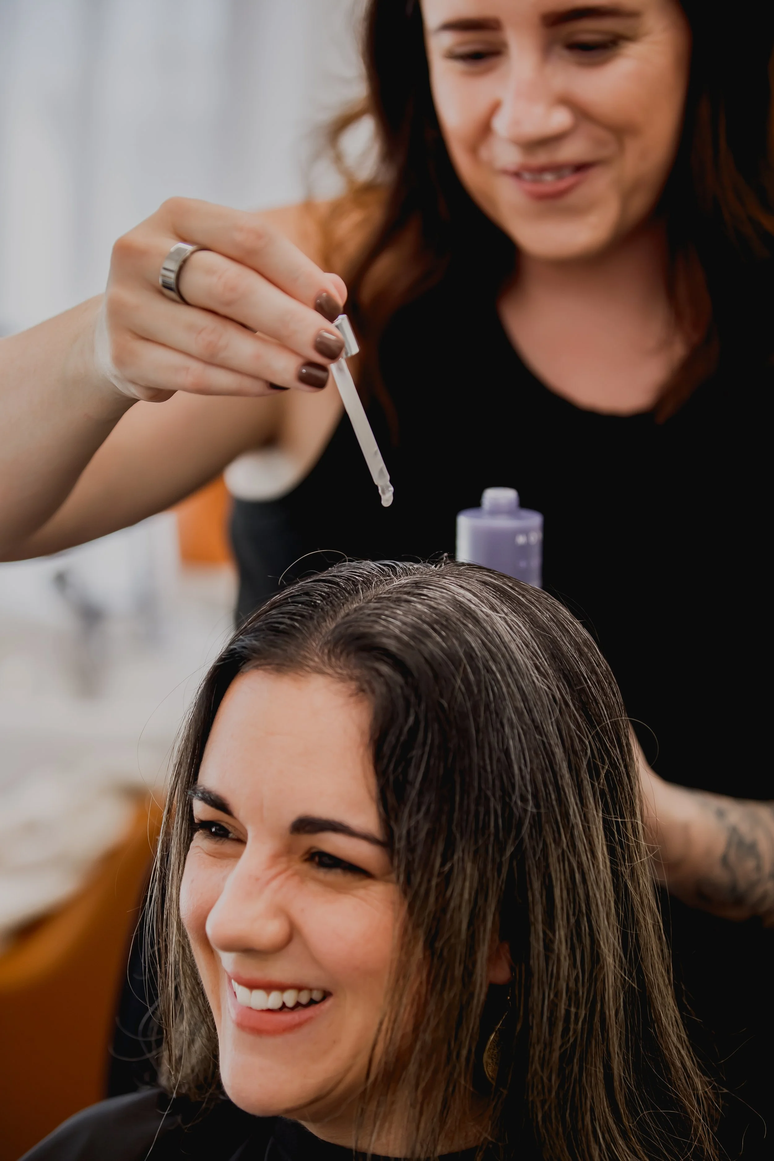 A woman with brown hair smiling and looking down, receiving a hair loss treatment involving a scalp product, while a hair loss specialist in the background applies it to her hair.