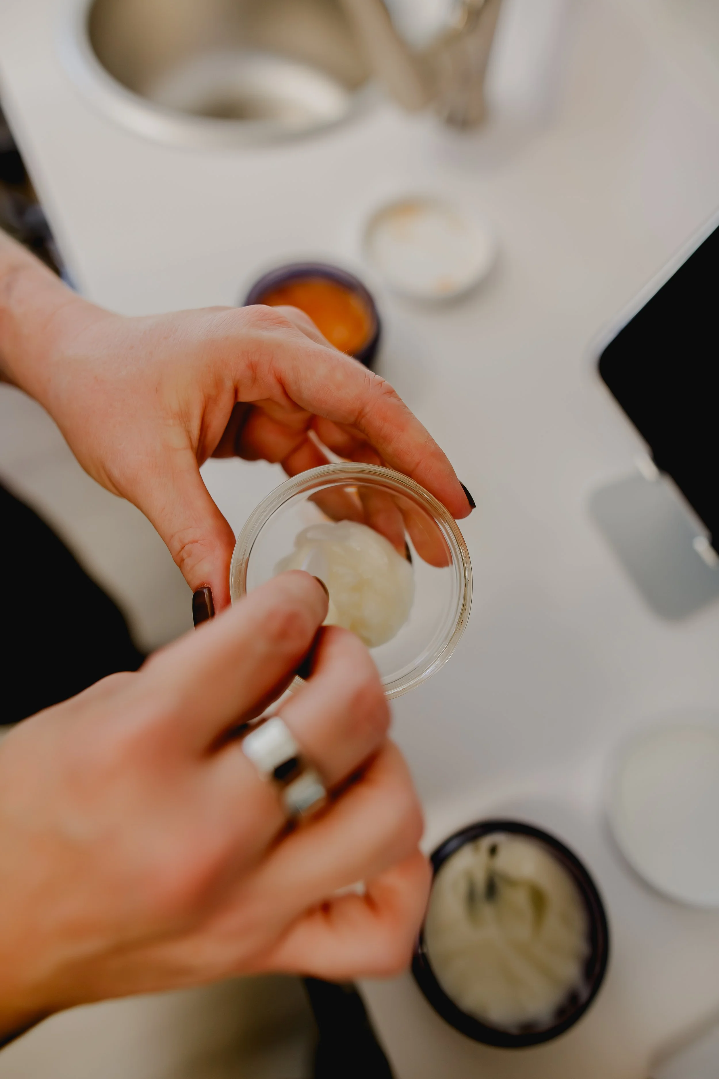 A person holding a small clear container with a white substance, with several open jars of similar contents on a white surface in the background.