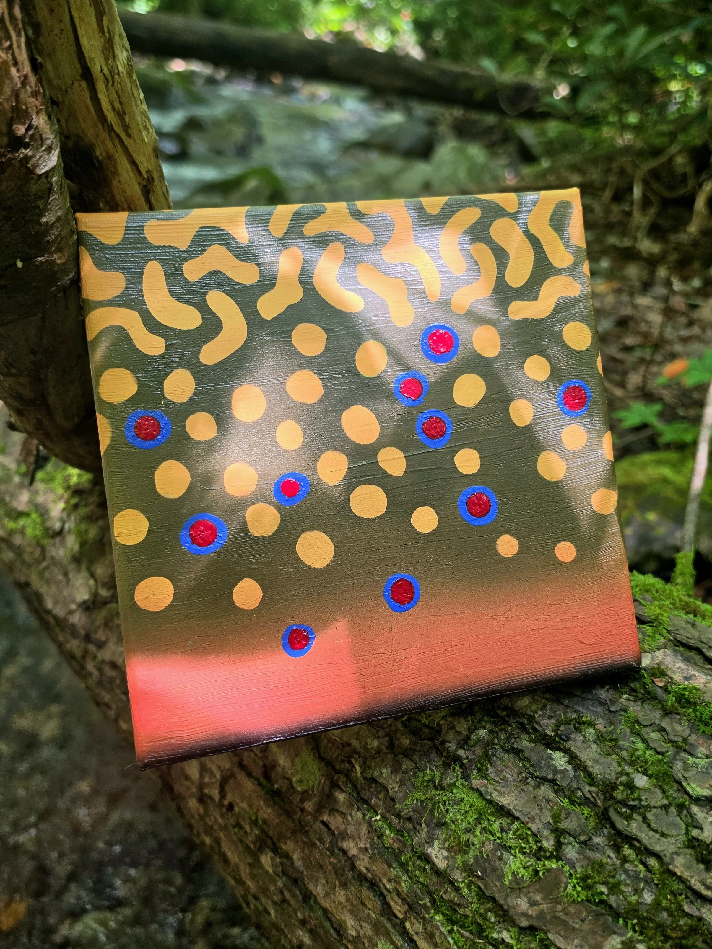 A colorful painting leaning against a tree trunk with green moss, featuring a pattern of yellow circles, black curvy lines, and red and blue dot accents on a metallic surface.