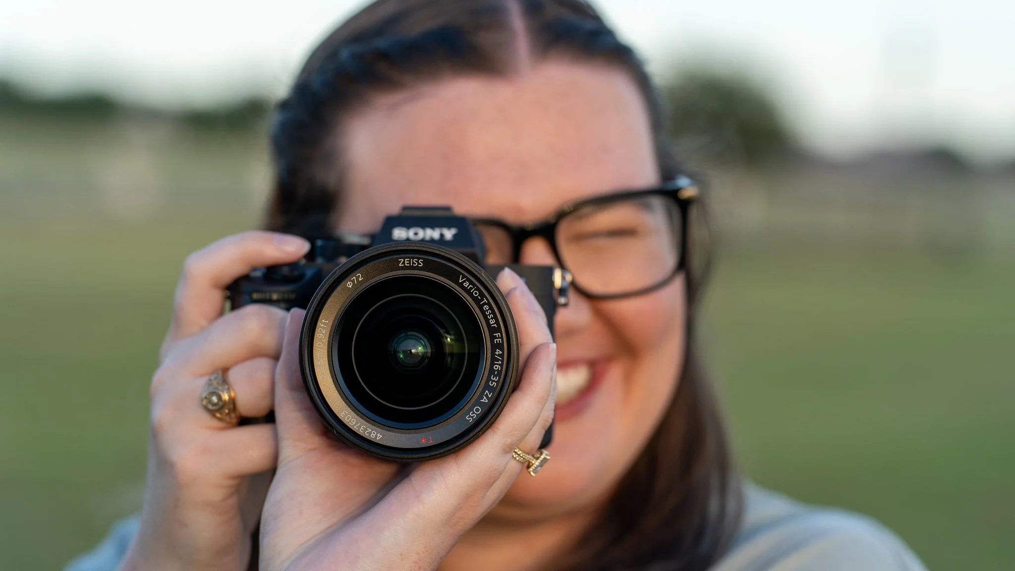 Person with long dark hair and glasses taking a photo with a Sony camera outdoors, smiling, with a blurry natural background.