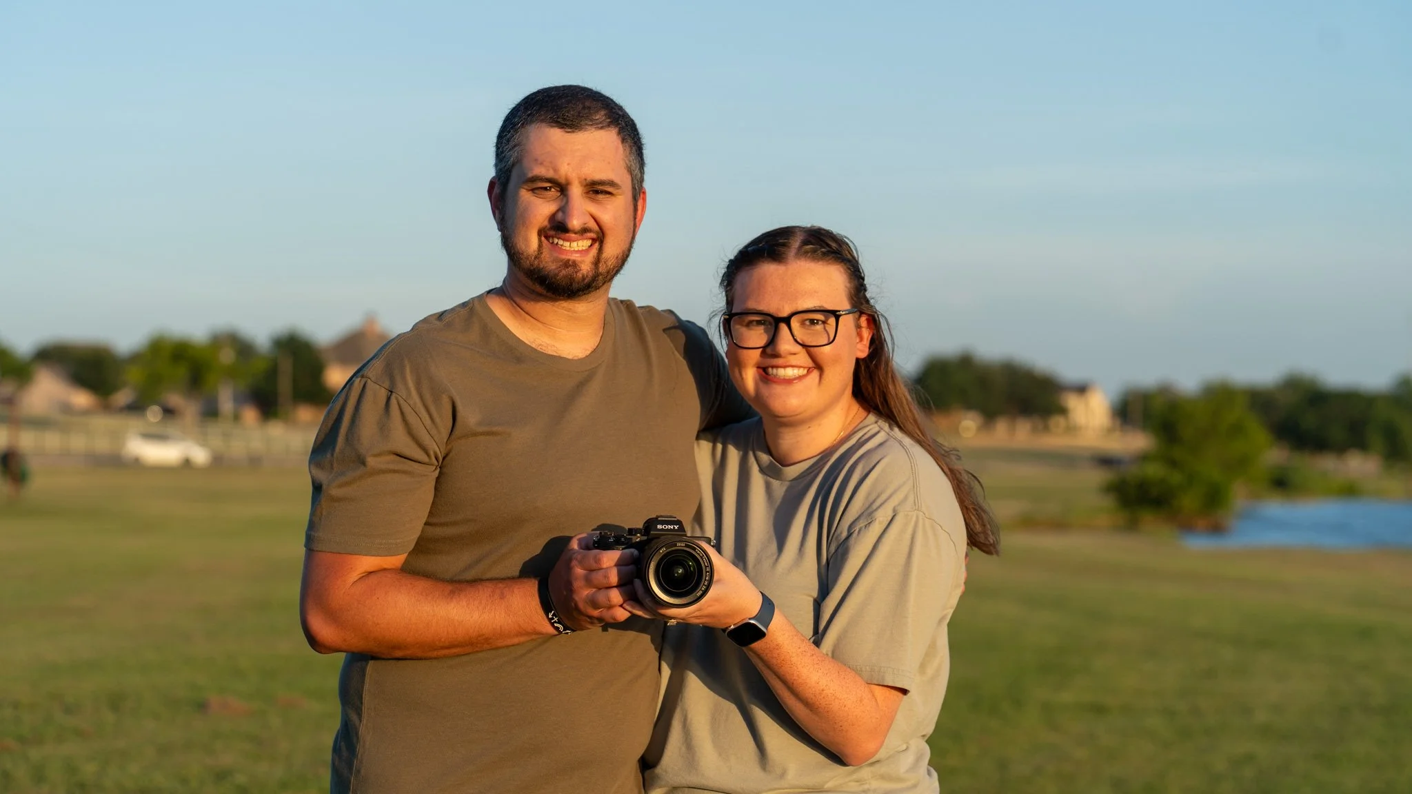 A man and woman stand outdoors in a grassy area, smiling at the camera during sunset, with trees, houses, and a body of water in the background.