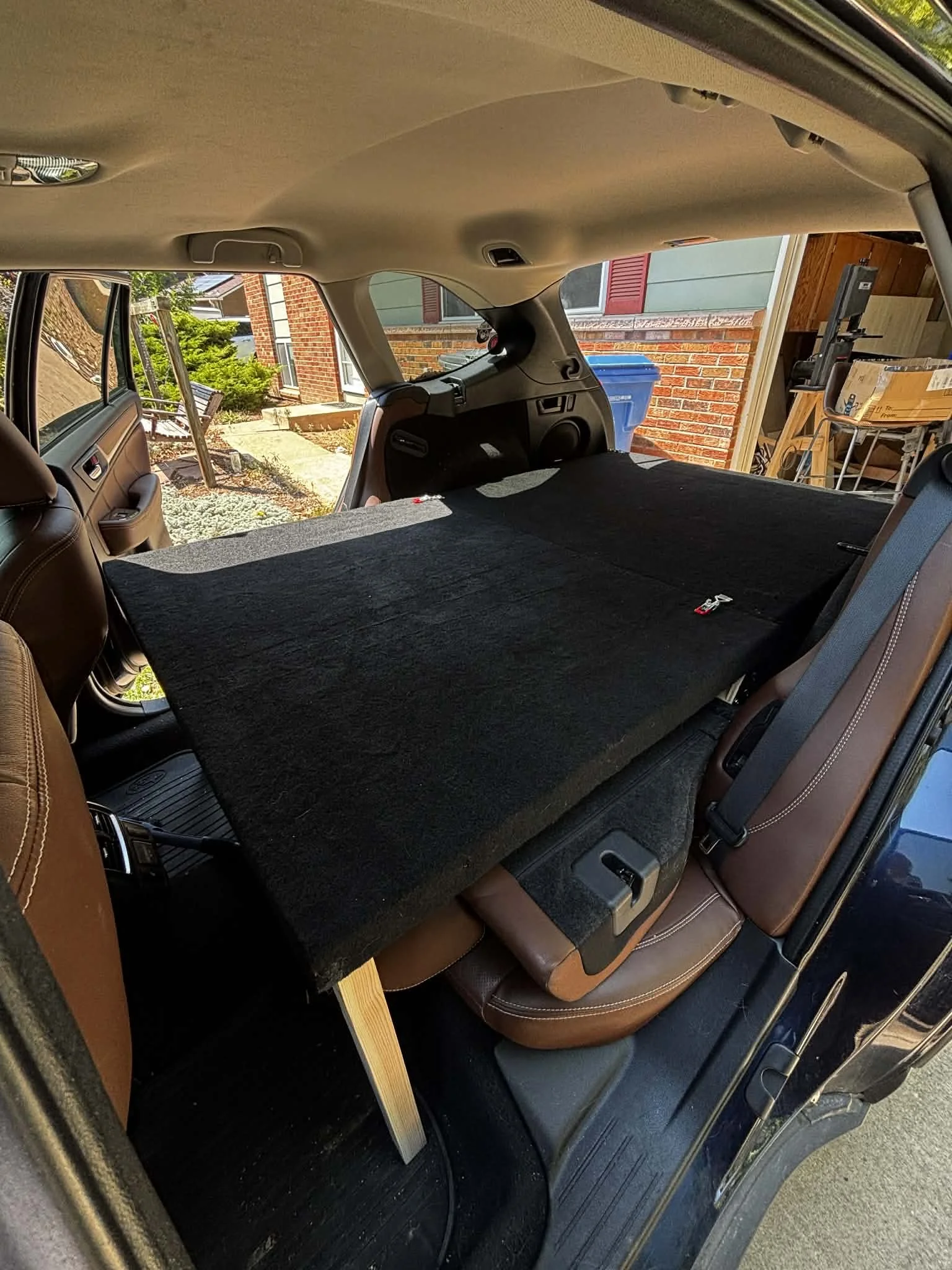 The back of an SUV with the rear seats folded down, creating a flat loading surface covered with a black mat. Outside the vehicle, a brick house with a window, a blue recycling bin, and green plants are visible.
