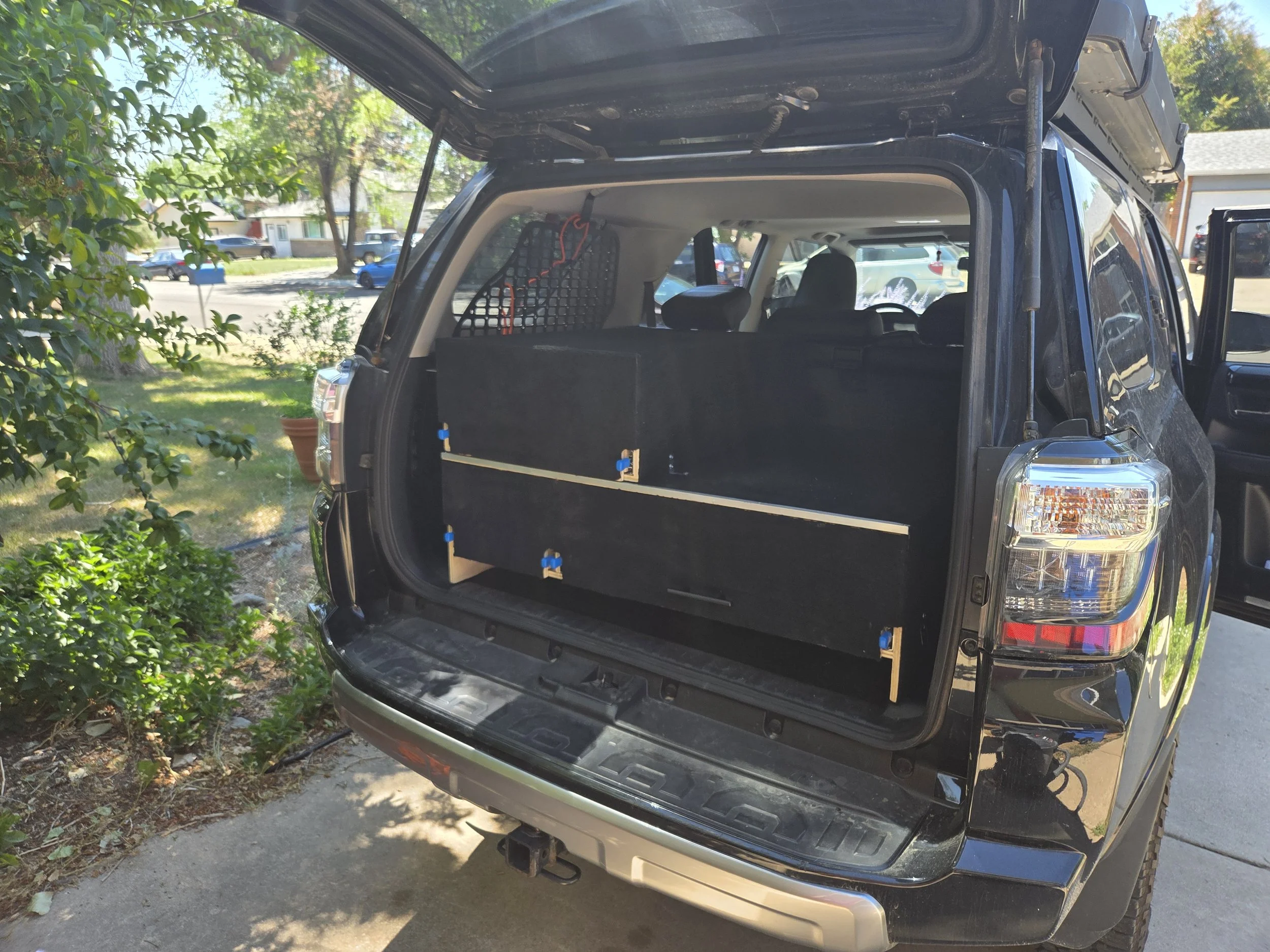The trunk of a black SUV open, revealing a flat black interior with some orange ties securing items inside, parked on a driveway next to some plants and a lawn with houses and other cars in the background.