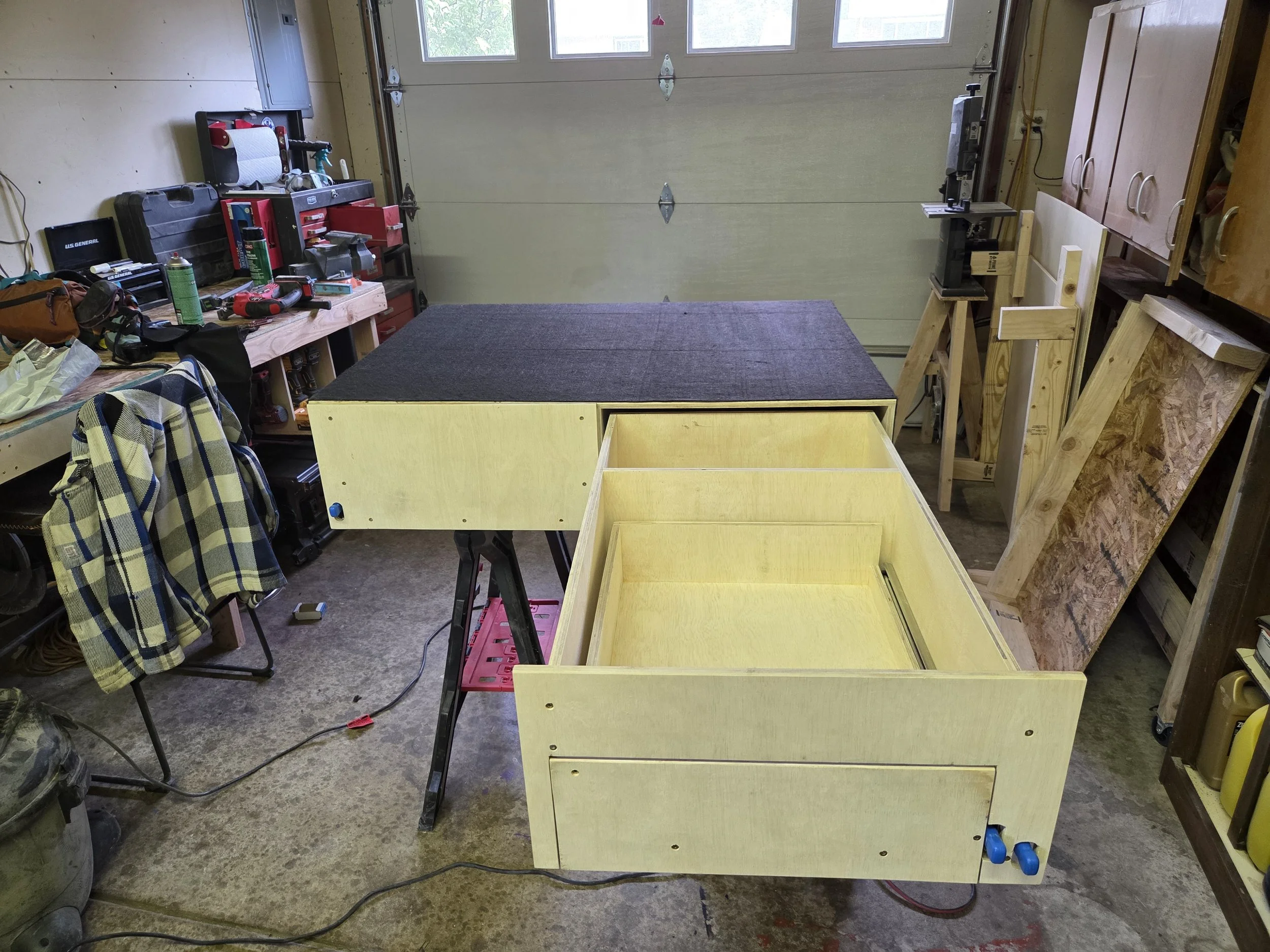 A cluttered garage workshop with a workbench, tools, and materials for woodworking and repairs, including a drill, tape, and wooden boards.