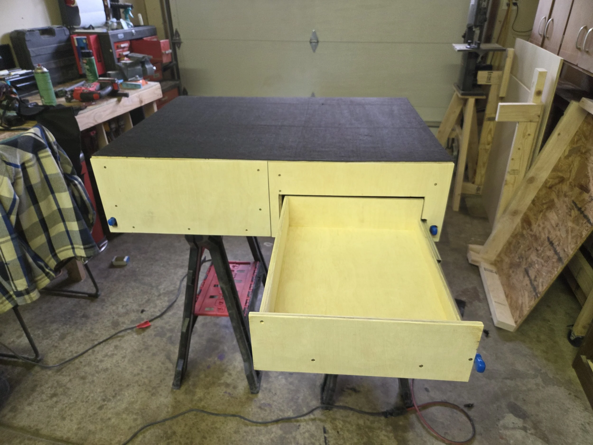 A partially assembled piece of furniture, possibly a desk or a cabinet, with yellow-painted wooden panels and black cushions on top, set up in a workshop or garage space with tools and wooden planks around.