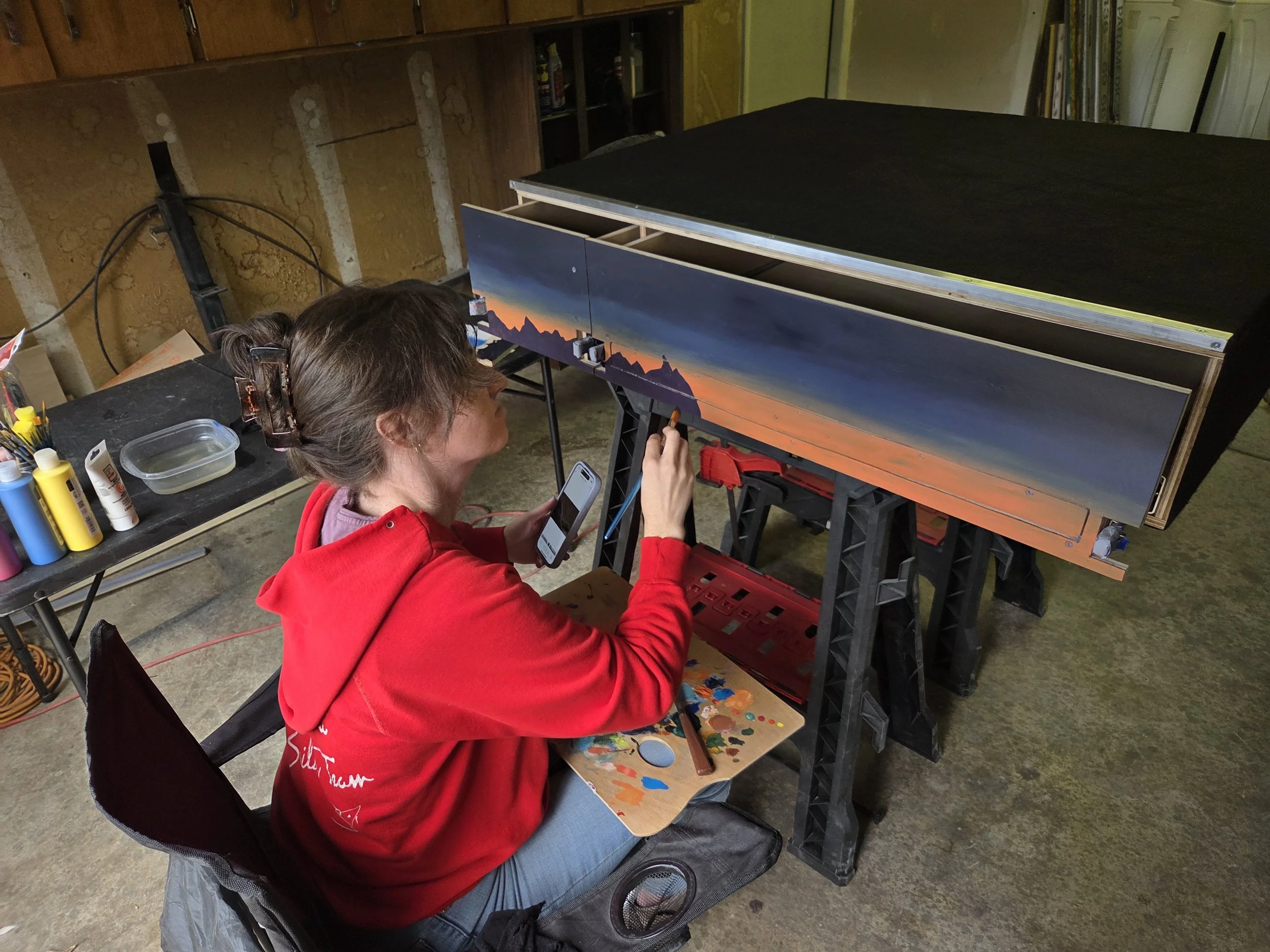 A woman in a red hoodie is painting a landscape mural on a large wooden panel in a workshop. She is seated, holding a paintbrush, with a palette of colors nearby. The mural depicts mountains and a sunset sky. The workshop has exposed wooden walls, various art supplies on a table, and an unfinished black canvas on top of the mural.