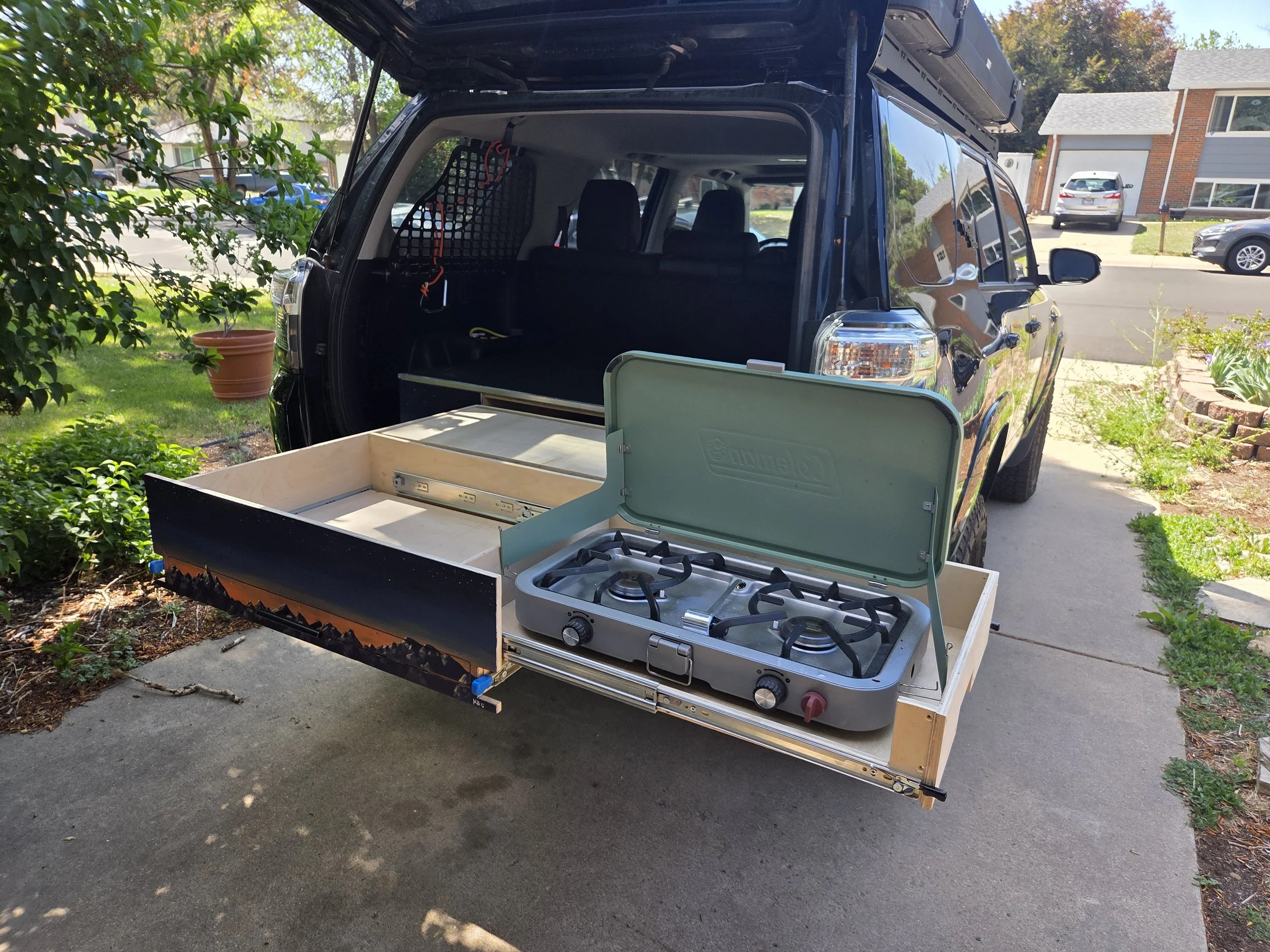 A black SUV with its rear hatch open, parked on a driveway with a kitchen stove placed partially outside. The area is residential with trees, plants, and other cars in the background.
