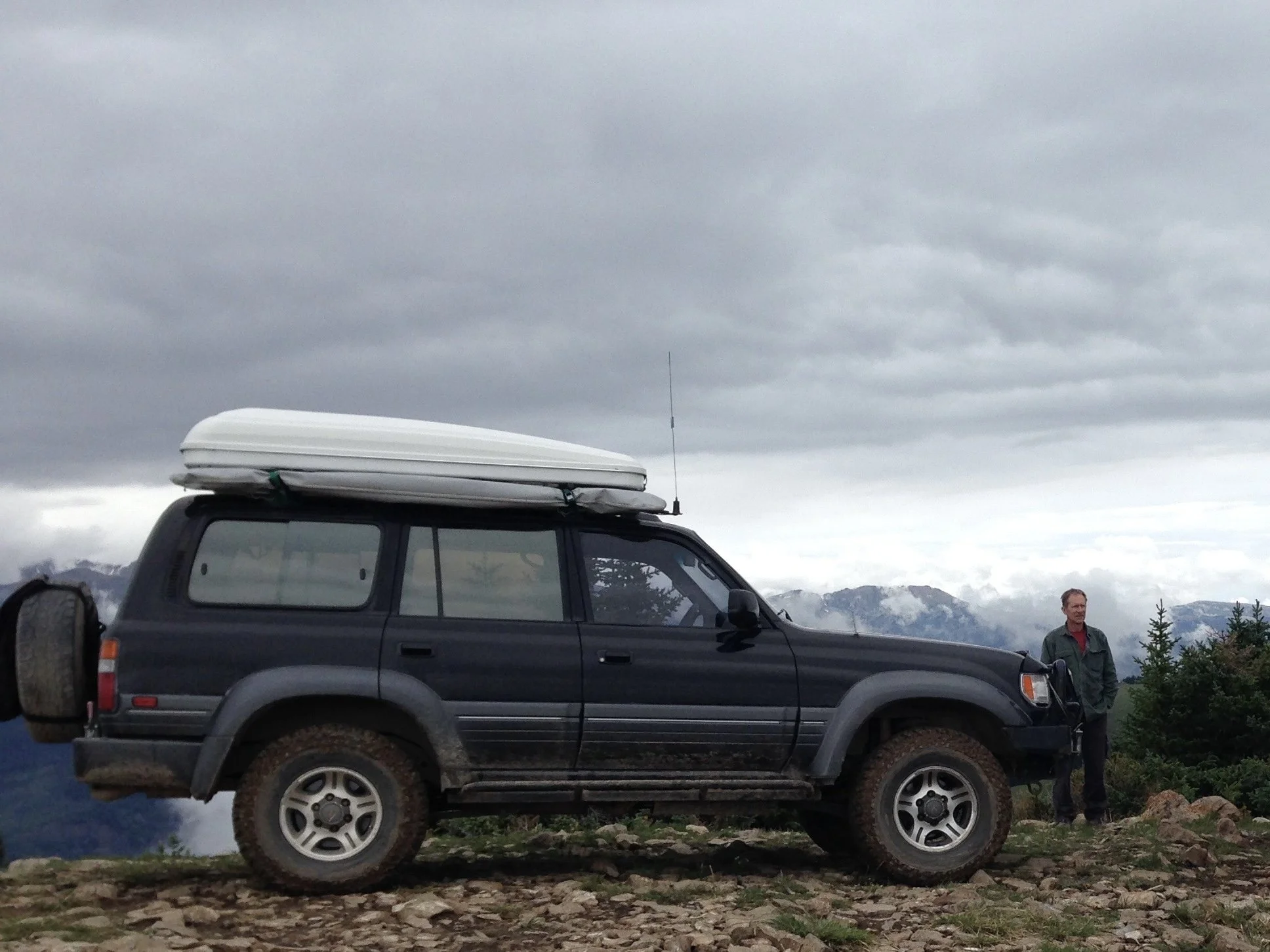 A black SUV with a roof cargo box parked on a rocky terrain with a mountainous background. A man in a green jacket stands near the rear of the vehicle under a cloudy sky.