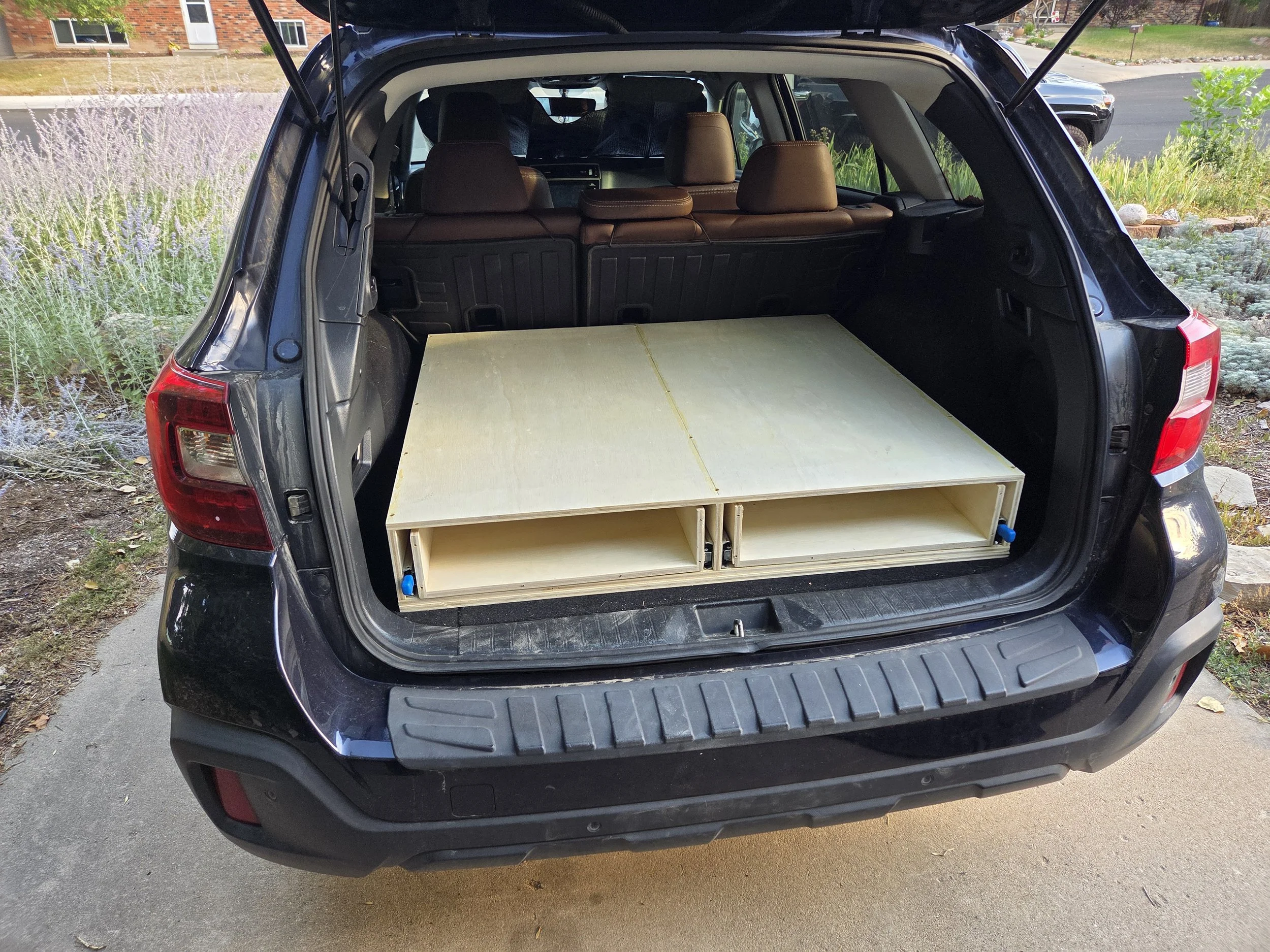 The open trunk of a black SUV with a custom wooden storage unit inside, parked on a residential street with houses, trees, and a driveway in the background.